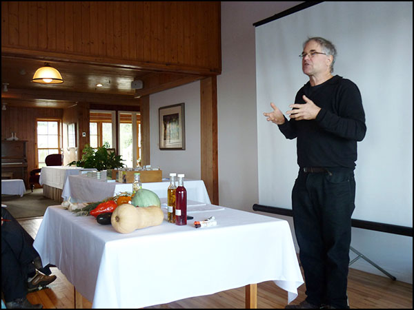 photo de formateur, vêtu de noir, cheveux courts blancs et lunettes, parant devant un écran blanc et une table sur laquelle il y a plusieurs légumes et vins.